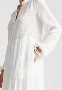 Robe blanche à manches longues en tissu texturé et rayé, avec un plastron boutonné à mi-hauteur et un design en plusieurs couches ; présente une coupe ample et des manches froncées.