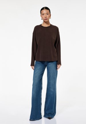 Kobieta o czarnych włosach, z złotymi kolczykami kołami, ubrana w luźną ciemnobrązową bluzkę z długim rękawem i szerokie niebieskie dżinsy, stojąca na białym tle.