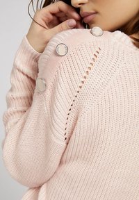 Pull en maille rose clair avec texture côtelée, orné de boutons décoratifs argentés sur l'épaule et présentant un léger motif ajouré.