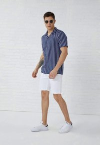 Uomo con occhiali da sole tondi, camicia a maniche corte a righe blu e bianche, pantaloni corti bianchi e scarpe da ginnastica bianche in piedi contro un muro di mattoni bianchi.