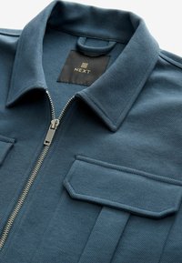 Jasje met kraag in een gestructureerde blauwe stof, voorzien van een ritssluiting aan de voorkant, twee klepzakken en een zwart label met "NEXT" in goud.