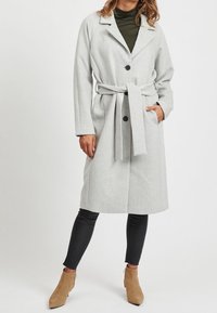 Lys grå uldblandet frakke med bælte, slået revers og to store knapper. Båret over en mørk rullekrave, parret med sorte leggings og beige ankelstøvler.