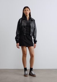 Μαύρο δερμάτινο κοντό μπουφάν με μακριά μανίκια, πάνω από μαύρο φόρεμα. Το μοντέλο φοράει μαύρες κάλτσες αστραγάλου και γκρι αθλητικά παπούτσια με κορδόνια και υφασμάτινες λεπτομέρειες.