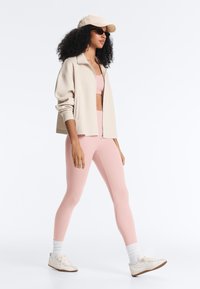 Woman with curly hair walking, wearing beige zip-up jacket, pink sports bra and leggings, white sneakers, socks, baseball cap, and sunglasses.