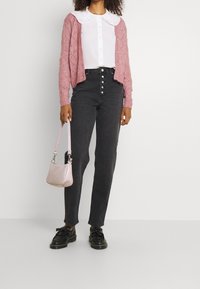 Žena v růžovém pleteném kardiganu, bílé blouze s kulatým límcem, černých džínách s vysokým pasem, černých mokasínech a držící malé růžové kabelky.