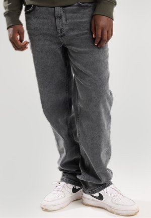 Osoba ubrana w ciemnoszare jeansy i białe buty Nike stojąca na jednolitym tle.