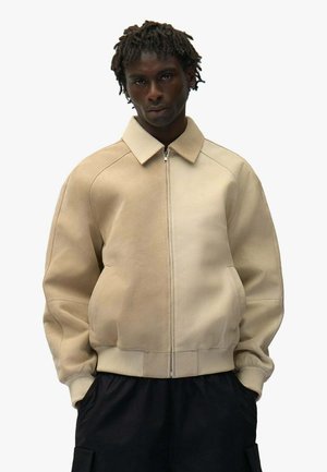 Uomo con dreadlocks corti che indossa una giacca beige bicolore con zip, polsini e collo a coste, in piedi con le mani nelle tasche.