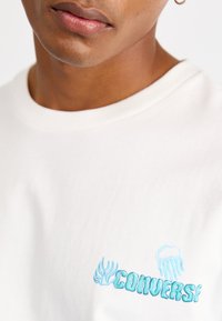 Crewneck t-skjorte i lys kremfarget bomull, med lys blå brodert "Converse" tekst og manetdesign på venstre bryst.