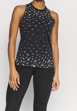 Vrouw draagt een zwart mouwloos sporttopje met kleine witte abstracte patronen en nauwsluitende zwarte leggings tegen een neutrale achtergrond.