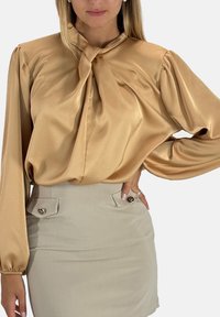 Blusa in raso beige chiaro con scollo annodato, maniche lunghe a sbuffo e texture liscia, abbinata a una gonna beige chiaro con bottoni accentuati.