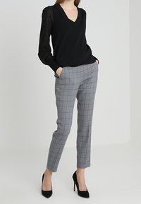 Musta neulottu V-aukkoinen neulepaita, jossa on teksturoidut mustat hihat, yhdistettynä harmaisiin ruutukuvioituihin räätälöityihin housuihin ja mustiin korkokenkiin.