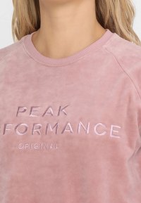Sudadera de forro polar de color rosa claro con cuello redondo, que presenta el texto bordado "PEAK PERFORMANCE ORIGINAL" en un rosa ligeramente más oscuro.