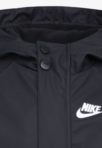 Čierna nylonová bunda s jemnou podšívkou, s dvoma čiernymi gombíkmi na golieri a bielym logom Nike na boku.