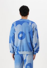 Blå sweatshirt med stora blommönster i ljusblått, med rund halsringning och ribbade ärmmuddar, visad bakifrån på en modell.