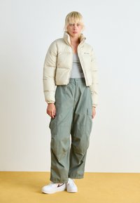 Veste en crème bouffante, haut gris, pantalon cargo vert et chaussures blanches avec des accents rouges. Style décontracté, coupe oversize et texture matelassée pour l'extérieur.