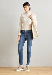 Beige csíkos, hosszú ujjú felső fodros dekoltázzsal, kék szűk farmerrel és világosszürke sportcipővel párosítva. A modellen egy krémszínű vászontáska van.