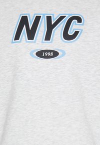 T-shirt in cotone grigio con bold lettering "NYC" in nero e azzurro chiaro, con "1998" in un ovale sottostante. Texture liscia, design casual.
