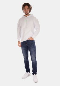 Felpa bianca in cotone con logo verde, abbinata a jeans in denim blu e sneakers bianche. Texture morbida, vestibilità casual e maniche lunghe.