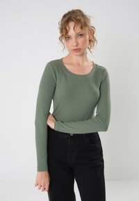 Maglietta a maniche lunghe in tessuto a coste verde oliva, con un ampio scollo. Abbinata a jeans neri a vita alta per uno stile a contrasto.
