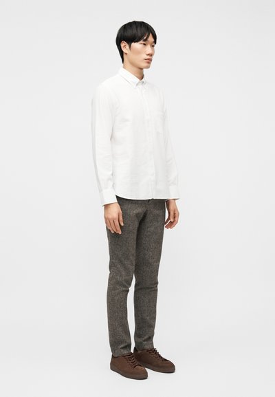 Νέος άνδρας που φορεί λευκό πουκάμισο με κουμπιά, γκρι υφασμάτινο παντελόνι και καφέ αθλητικά παπούτσια, στέκεται μπροστά σε απλό λευκό φόντο.