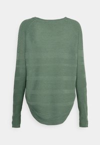 Maglietta a maniche lunghe verde con texture a coste, orlo arrotondato e vestibilità rilassata. Il tessuto appare morbido e leggero.