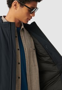 Giacca nera esterna con una texture liscia, indossata sopra una camicia a quadri marrone e un maglione blu, che mostra una tasca con zip all'interno della giacca.