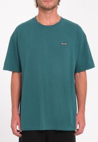 Homme portant un t-shirt à manches courtes teal avec un petit logo noir et blanc "Volcom" sur la poitrine gauche, se tenant devant un fond blanc uni.