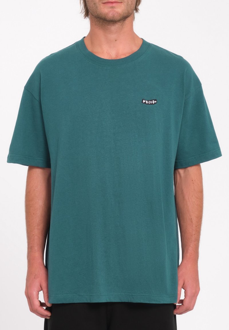 Homme portant un t-shirt à manches courtes teal avec un petit logo noir et blanc "Volcom" sur la poitrine gauche, se tenant devant un fond blanc uni.