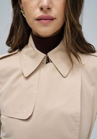 Manteau beige avec un grand col et une poche poitrine, associé à un haut marron foncé à col montant. Dispose d'une fermeture éclair au niveau du col.