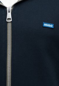 Κοντινή λήψη μαύρου ένδυματος με φερμουάρ και ασημένια φερμουάρ, με μπλε ορθογώνιο διακριτικό με το λογότυπο "HUGO" στο στήθος.