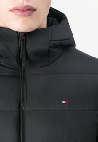 Jachetă puffer neagră cu glugă, cu închidere tip fermoar, având un mic logo de marcă în roșu, alb și albastru pe partea stângă a pieptului. Textură netedă.