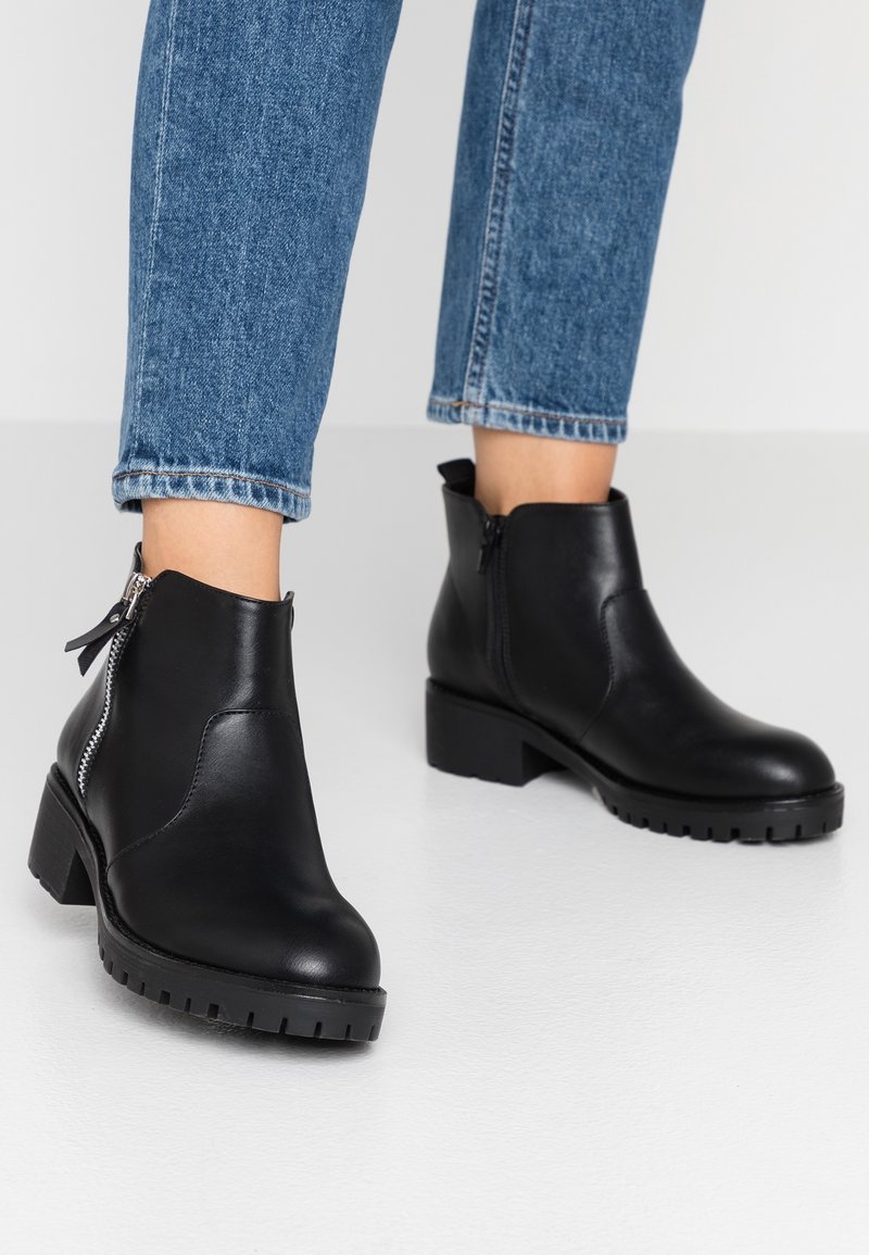 Personne portant des bottines en cuir noir avec des semelles épaisses et des fermetures éclair, associées à un jean en denim bleu sur un fond uni.