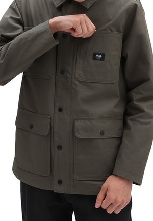 Personne portant une veste boutonnée vert olive avec trois poches avant, une poche poitrine étiquetée "VANS" et un pantalon noir.