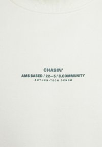 Bijela dukserica sa zelenim printanim tekstom: "CHASIN' AMS BASED / 22-5 / C.COMMUNITY AUTHEN-TECH DENIM." Okrugli izrez, mekana tekstura.