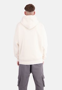 Cream-colored oversized hoodie with a large hood, ribbed cuffs, and hem. Worn over gray cargo pants with side pockets.