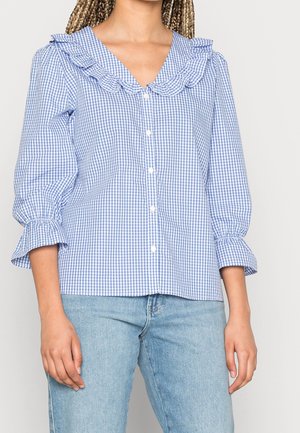 Kvinde iført blå og hvid ternet skjorte med knapper, flæsekrave og ærmer, kombineret med lyseblå jeans.