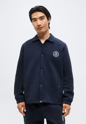 Homme portant une veste boutonnée bleu marine et un pantalon assorti avec un logo blanc circulaire sur la poitrine gauche et la cuisse, sur fond gris uni.