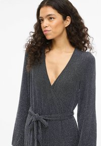 Tamsiai pilka su subtiliu metaliniu blizgesiu, ilgomis rankovėmis ir giliais V formos iškirpte, su derančiu audinio diržu, surištu juosmens srityje.