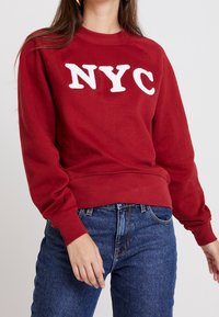 Red sweatshirt made of soft fabric featuring white embroidered "NYC" letters on the front, paired with blue denim jeans.