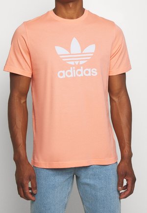 Brzoskwiniowy bawełniany t-shirt z białym logo Adidas z trzema paskami i motywem liści, okrągłym dekoltem i krótkimi rękawami.