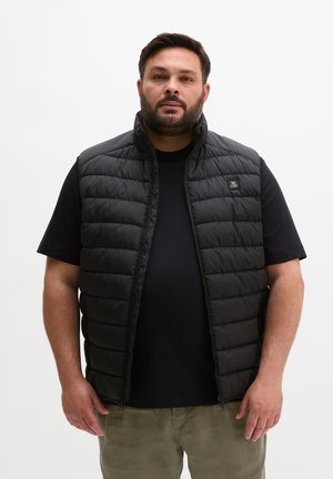 Homme aux cheveux courts et foncés et barbe, portant un gilet matelassé noir sur un t-shirt noir et un pantalon vert olive, debout devant un fond blanc uni.