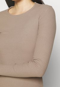 Haut à manches longues beige en tissu côtelé avec un col rond et des manches ajustées, présentant une texture lisse et un léger élasticité.