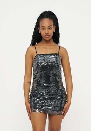 Femme aux longs cheveux tressés portant une mini-robe noire à paillettes avec des bretelles fines, debout devant un fond clair uni.