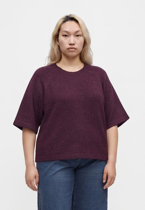 Ung kvinde med langt blond hår iført en løs plommefarvet kortærmet sweater og blå jeans, der står mod en ensfarvet baggrund.