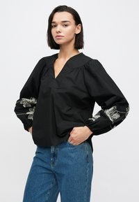 Čierna blúzka s hlbokým výstrihom do tvaru V a naberanými rukávmi, zdobená bielou kvetinovou výšivkou na rukávoch. Materiál pôsobí hladko a ľahko.