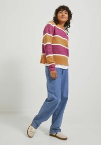 Sweter w paski w różowych, pomarańczowych i białych włóknach, charakteryzujący się luźnym krojem i długimi rękawami, zestawiony z jasnoniebieskimi dżinsami i jasnymi butami.