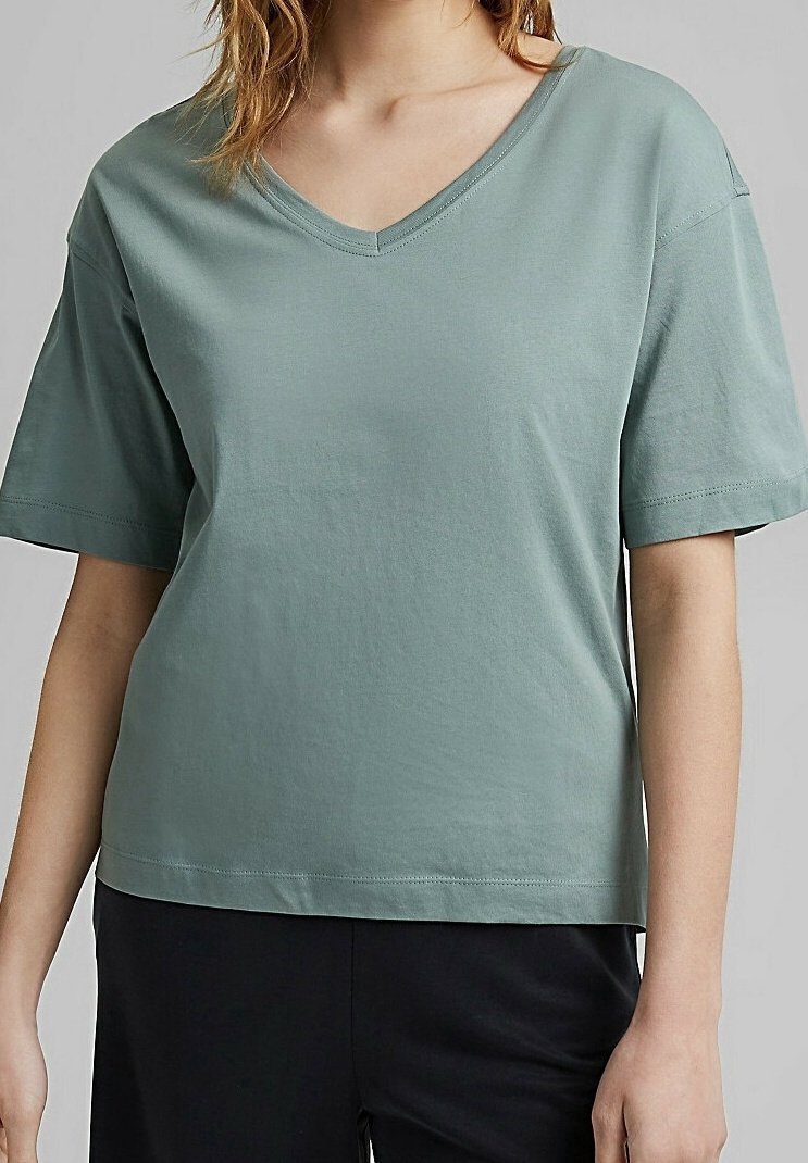 Femme portant un t-shirt ample à manches courtes de couleur vert sauge avec un col en V, associé à un pantalon noir sur un fond uni.