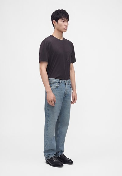 Jeune homme debout portant un t-shirt noir à manches courtes, un jean bleu clair et des chaussures habillées noires sur un fond clair uni.