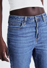 Jean en denim bleu au design ajusté, présentant un style classique à cinq poches et des coutures jaunes contrastantes aux coutures.
