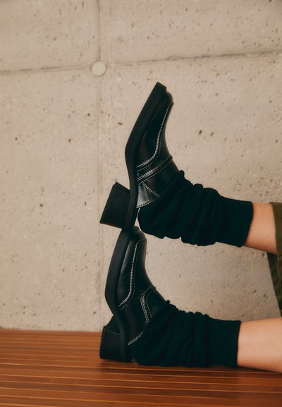 Botas de tobillo de cuero negro con costuras blancas, tacones de bloque y calcetines negros fruncidos, descansando sobre una superficie de madera contra una pared de concreto.